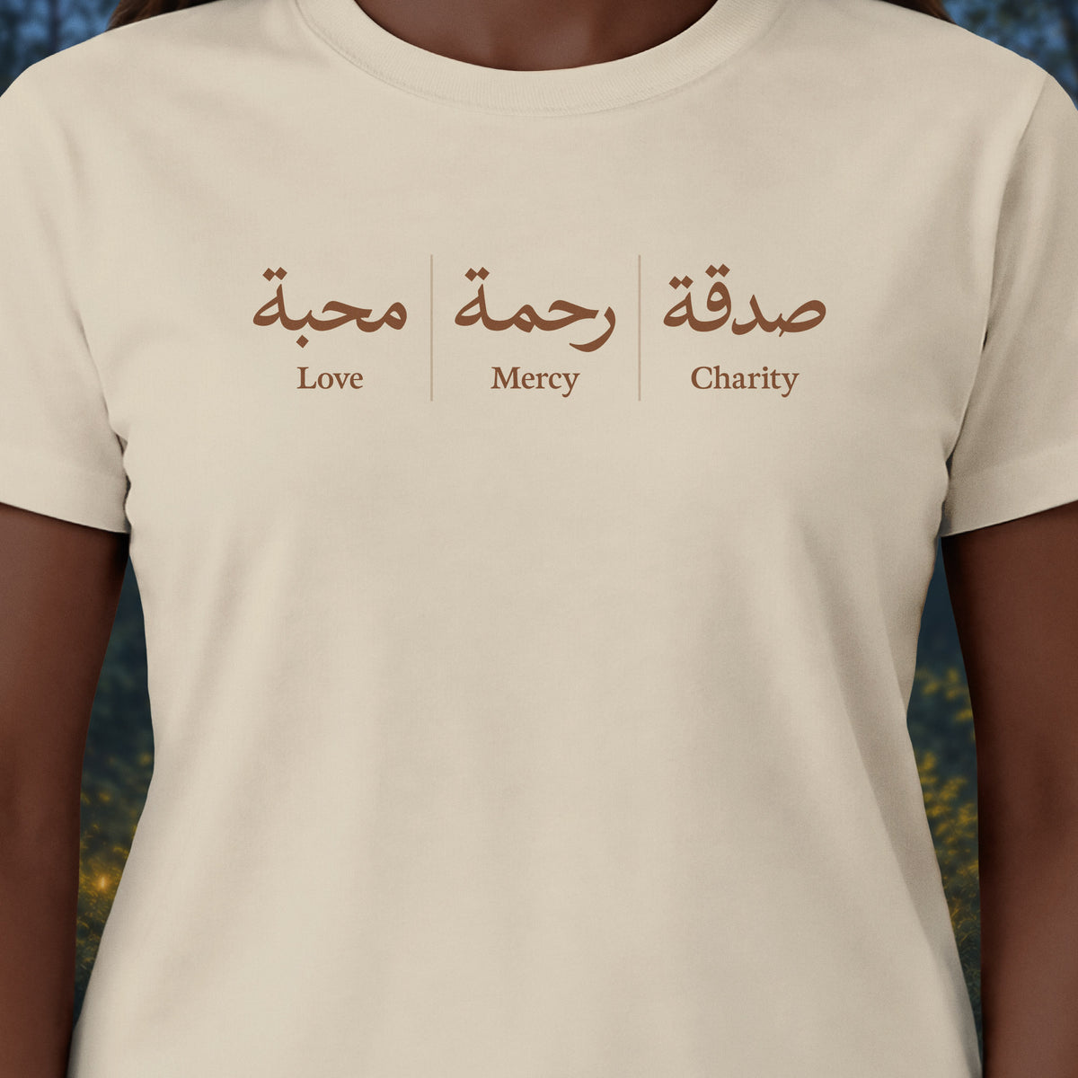 A close-up of a person wearing a cream-colored t-shirt with a brown graphic print on the chest. The print features Arabic calligraphy and the English words 'Love', 'Mercy', and 'Charity' underneath.