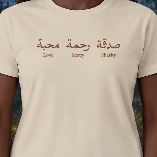 A close-up of a person wearing a cream-colored t-shirt with a brown graphic print on the chest. The print features Arabic calligraphy and the English words 'Love', 'Mercy', and 'Charity' underneath.